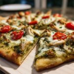 Chicken pesto flatbread topped with melted mozzarella, tender chicken chunks, and swirls of basil pesto, garnished with fresh basil leaves and cherry tomatoes, served on a white plate in a home kitchen setting.