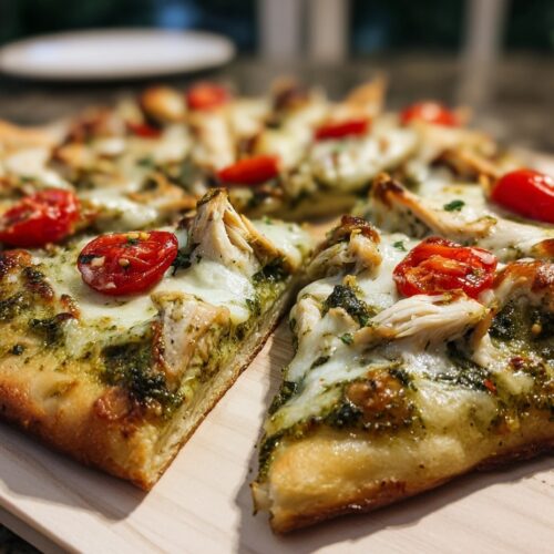 Chicken pesto flatbread topped with melted mozzarella, tender chicken chunks, and swirls of basil pesto, garnished with fresh basil leaves and cherry tomatoes, served on a white plate in a home kitchen setting.