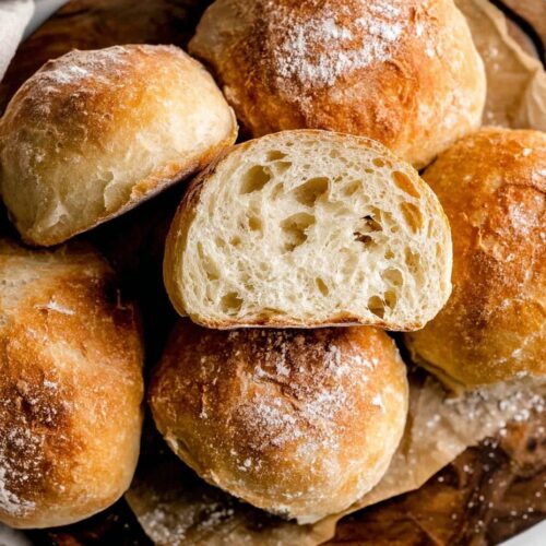 Golden-brown homemade ciabatta rolls in rustic wicker basket with linen towels, showing flour-dusted crusty surfaces and authentic artisan bread texture
