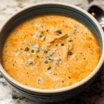 Creamy buffalo chicken soup in white bowl with tender chicken, Frank's Red Hot sauce, blue cheese crumbles, fresh scallions, carrots, and celery on wooden board