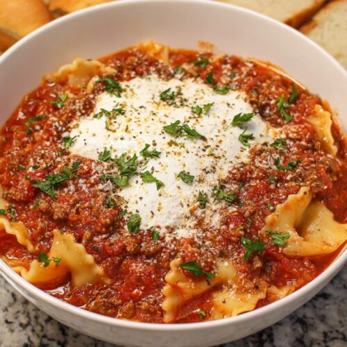 Hearty crockpot lasagna soup in white bowl with ground beef, Italian sausage, tender pasta, melted three-cheese topping, and fresh parsley on wooden board