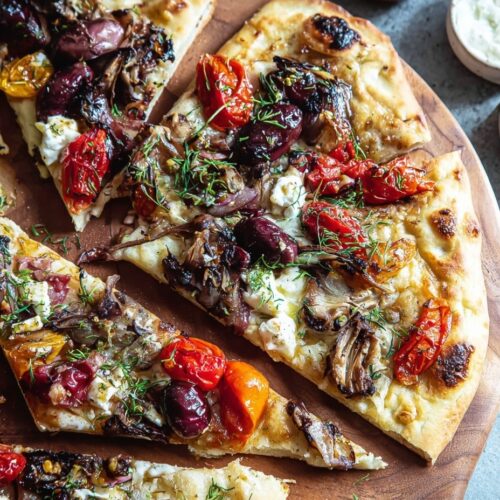 Easy Mediterranean flatbread on parchment paper topped with roasted red peppers, artichokes, kalamata olives, red onion, feta cheese, mozzarella, fresh parsley, and balsamic glaze drizzle