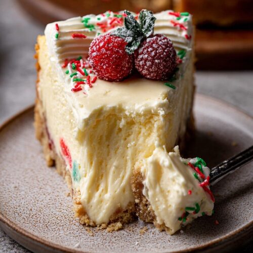 Slice of sugar cookie cheesecake showing layers of cookie crust, cream cheesecake with cookie dough balls, white chocolate ganache and festive sprinkles